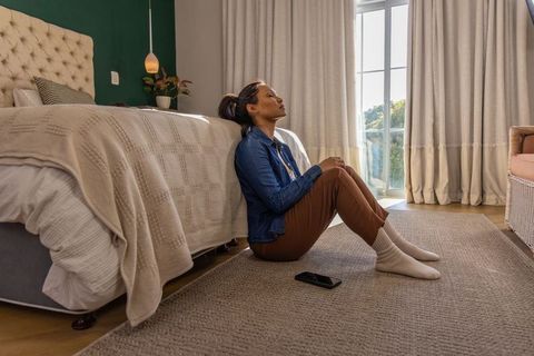 Asian woman in 30s sitting on bedroom floor leaning against tufted bed near window
