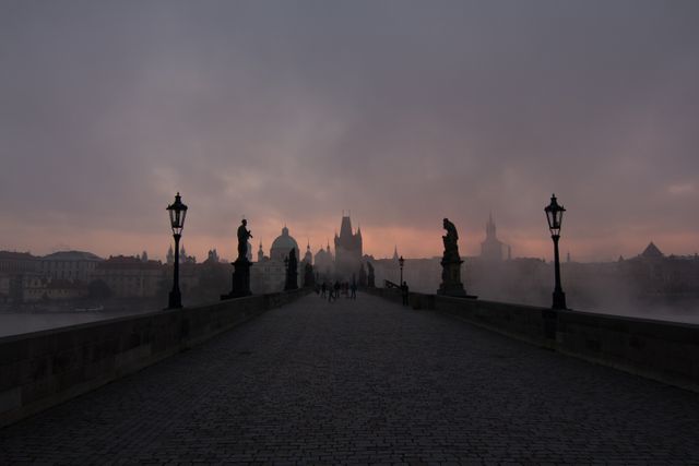 Image shows an old bridge shrouded in fog at dawn. Silhouetted statues and lamp posts line the bridge as the cityscape emerges in the background. This image is ideal for use in travel brochures, city guides, and articles about historical European architecture. It evokes a sense of mystery and historical allure, perfect for storytelling and introducing themes of history and travel.