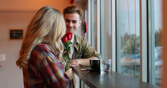 Two young adults sharing a romantic moment in a coffee shop by a large window. The woman is holding red roses while the man smiles at her. Perfect for themes like romance, relationships, dating scenes, and special moments. Ideal for use in Valentine's Day promotions, romantic articles, or greeting cards.