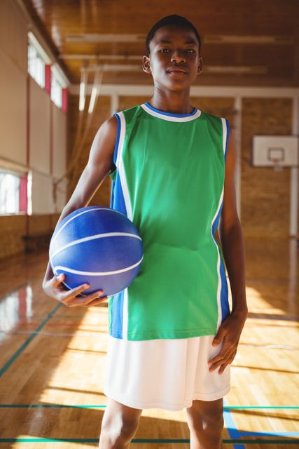 Young basketball player standing confidently in gym holding ball. Ideal for sports training programs, youth athletic promotions, team spirit campaigns, and fitness-related content.