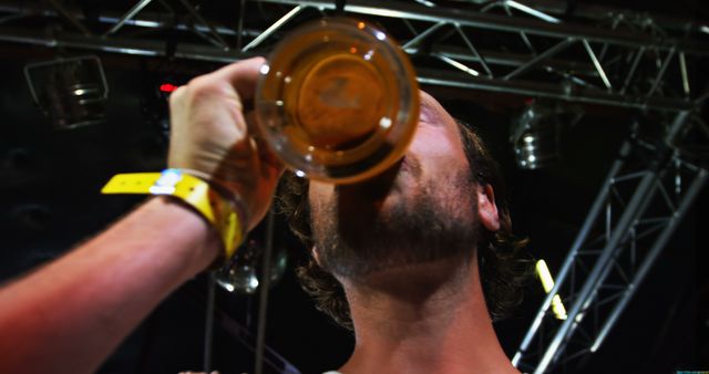 Man drinking beer from glass at night music festival. Stage lights and metal structures overhead suggest presence of live music or entertainment. Ideal for use in media related to nightlife, music festivals, social events, or beer industry.