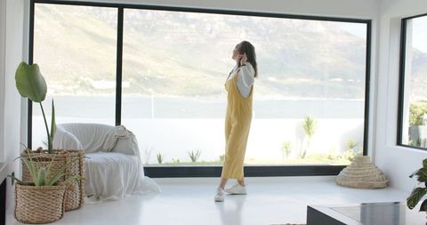 Woman gazing through floor-to-ceiling windows in sunlit coastal living room with greenery