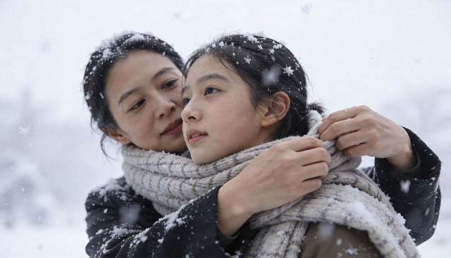 Mother wrapping daughter in ribbed knit scarf during gentle snowfall in winter park