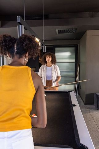 Black woman and mixed-race friend playing pool in modern game room, casual summer leisure