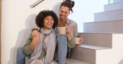 Laughing couple sipping coffee on modern tiled staircase, cozy minimalist interior