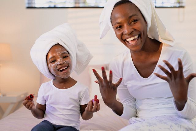 Mother and daughter are enjoying a spa day at home, wearing towels on their heads and painting nails. This image is perfect for promoting family bonding, self-care routines, and beauty products. It can be used in advertisements, blogs, and social media posts about parenting, relaxation, and home activities.