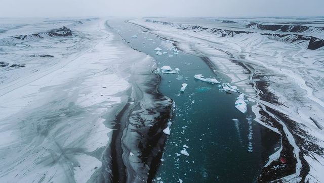 Winding glacier channel cutting through snow-dusted polar fjord with ice floes and red buoy
