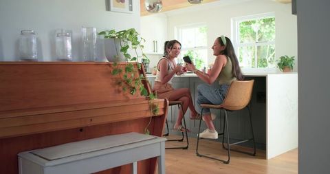 Two women laughing and sharing drinks at modern kitchen island near piano