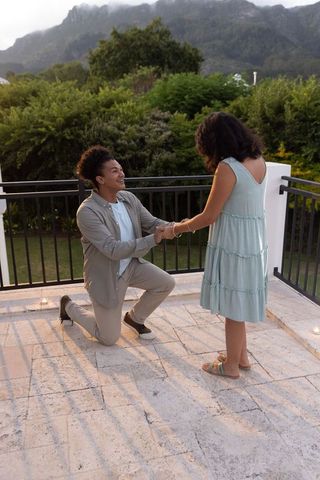 Rooftop proposal at dusk — diverse couple kneeling and holding hands by candlelight