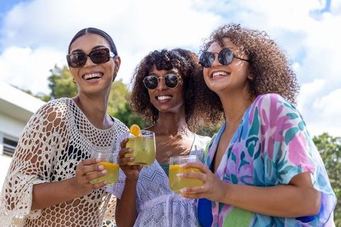 Smiling diverse women toasting citrus cocktails poolside wearing sunglasses summer celebration