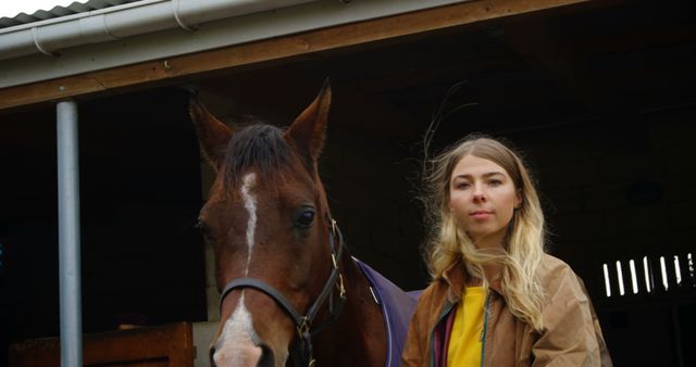 A young blonde woman stands beside a brown horse in a stable, looking directly at the camera. Her casual attire suggests an equestrian enthusiast or professional horse caretaker. Suitable for use in topics related to horse riding, equestrian sports, rural life, animal care, agriculture, or countryside living. Ideal for websites, blogs, and promotional materials for equestrian centers, farms, and rural tourism.