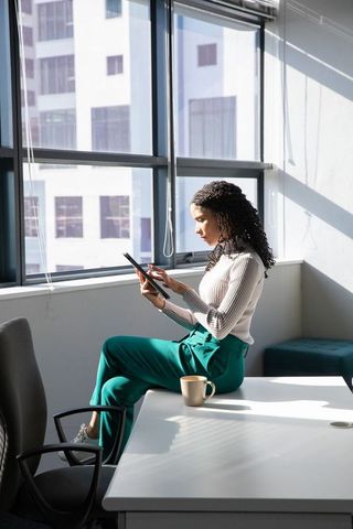African american woman sitting on desk tapping tablet in sunlit minimalist office