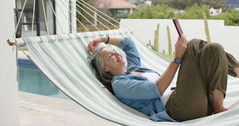 Mature woman relaxing on hammock by pool using smartphone in denim shirt and smartwatch