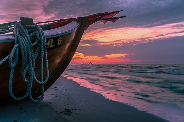 Wooden boat with ropes resting on sandy beach at sunset. Serene scene with colorful sky reflecting on calm ocean waters. Ideal for travel agencies, nature-related content, relaxation themes, and motivational quotes.