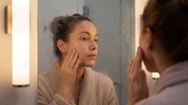 Woman applying facial cleanser at fogged bathroom mirror during evening skincare routine
