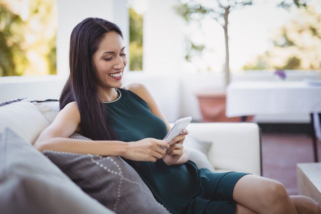 Woman sitting on a comfortable sofa in a restaurant, using her mobile phone and smiling. Ideal for themes related to modern lifestyle, technology, communication, leisure, and social media. Perfect for advertisements, blog posts, and articles about connectivity, relaxation, and contemporary living.