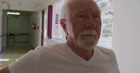Reflective senior man standing in sunlit care home corridor wearing white v-neck tee gazing ahead
