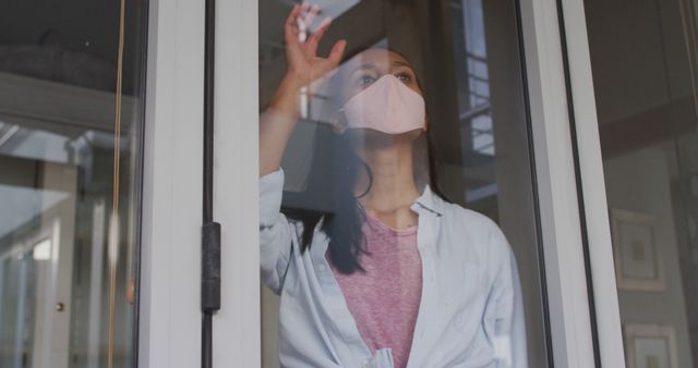 Woman wearing a face mask looking through window, contemplating. Could be used for content related to pandemic, mental health, isolation, or protection during quarantine.