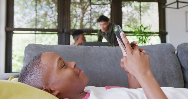 Boy lying on couch using smartphone casually in living room. Suitable for themes on youth, technology, leisure activities, and communication. Can be used in advertisements for smartphones, apps, or internet services, as well as content related to digital lifestyle and modern living.