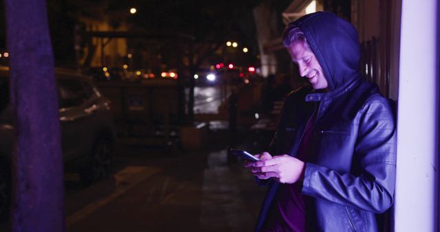 Young man in a hoodie is leaning against a wall while using smartphone on a city street at night. His face is illuminated by the phone screen, and he is laughing while using the device. Perfect for themes related to modern lifestyles, urban nightlife, youth culture, and digital communication.