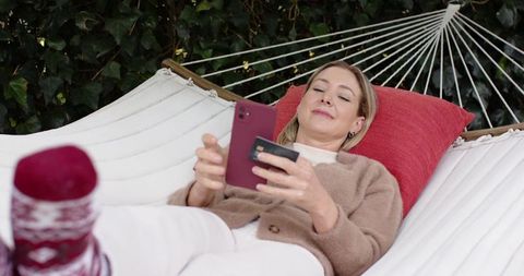 Relaxing woman using smartphone and credit card on hammock in backyard with red pillow