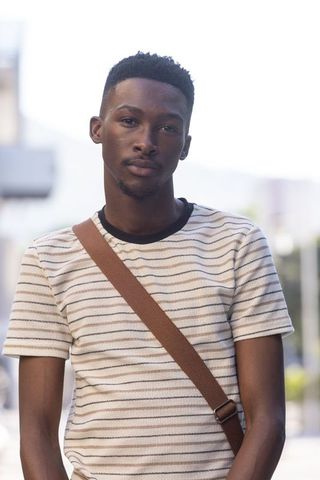 Young man standing on urban sidewalk wearing striped knit shirt and brown crossbody strap