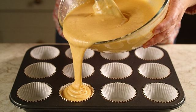 Pouring cupcake batter into paper liners closeup holding glass bowl spatula baking