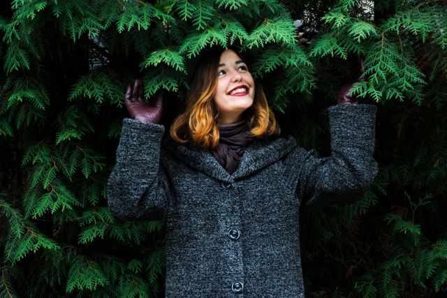Woman is delighted while standing near an evergreen tree, wearing a winter coat and brown gloves. Good for showcasing winter fashion, outdoor activities, seasonal joy, nature appreciation, or promotional material for autumn and winter clothing collections.