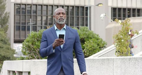 Mature businessman walking through urban plaza holding smartphone, looking focused