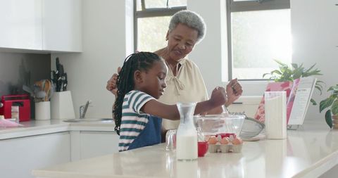 Senior african american grandmother teaching granddaughter to crack eggs while baking in bright kitc