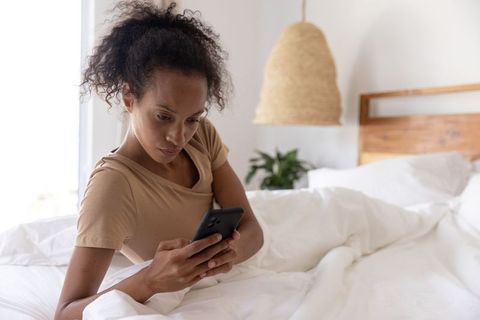 African american woman relaxing in bed scrolling smartphone under white duvet in bright bedroom