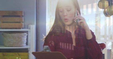 Retail counter staff holding clipboard and talking on smartphone with sunlit window glare