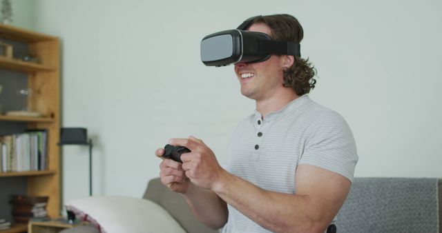Man engaging in virtual reality gaming, holding game controller with both hands, sitting on gray couch in casual home interior with wooden bookshelf in background. Useful for depicting modern entertainment, gaming technology, and virtual reality experience in advertisements, blogs, and social media.