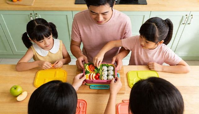 Father and daughters preparing colorful bento lunchboxes together on kitchen table