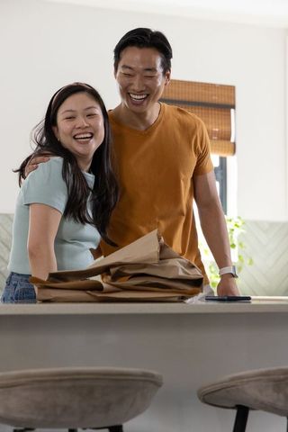 Asian couple laughing at kitchen island unpacking groceries and checking smartphone