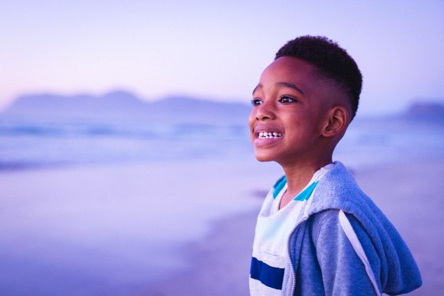 Side view of smiling african american boy looking away while standing at beach against clear sky. copy space, sunset, childhood, nature, unaltered, lifestyle, enjoyment and holiday concept.