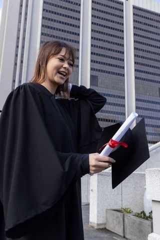 Asian graduate smiling while holding red-ribbon diploma and mortarboard amid city skyline