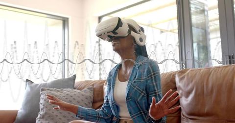Woman wearing VR headset reaching on sofa, enjoying immersive virtual reality at home