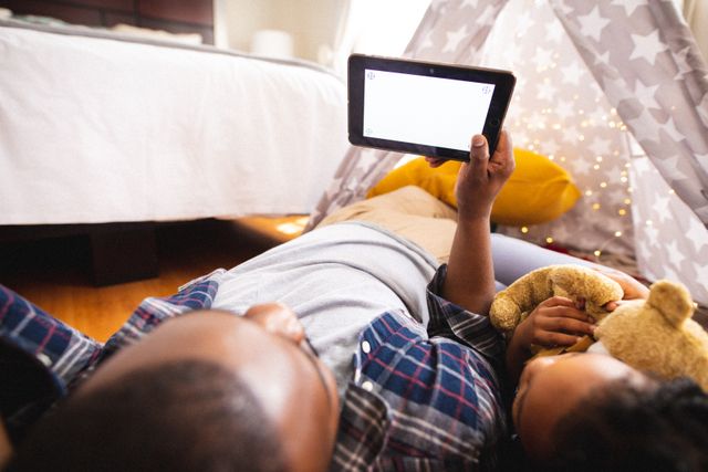 African american father sharing digital tablet with daughter lying in tent at home. unaltered, family, togetherness and technology concept.