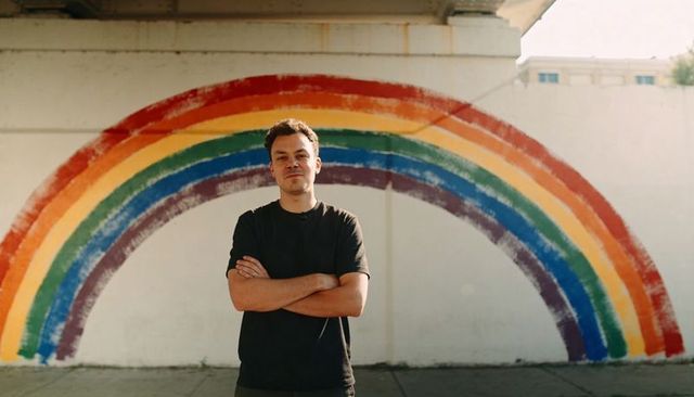 Confident man standing before rainbow mural on urban wall, pride support and street style