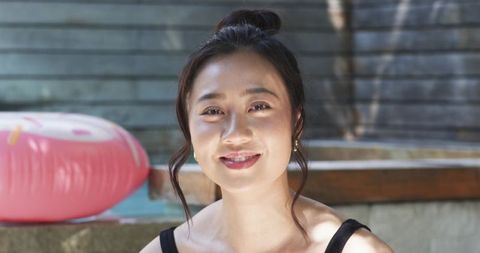 Young Asian woman smiling at backyard poolside wearing black tank top and hoop earrings