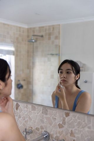 Young asian woman examining skin in bathroom mirror applying cotton pad