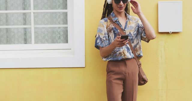 Woman standing by yellow wall wearing summer outfit, listening to music with headphones, using smartphone. Excellent for fashion blogs, digital lifestyle promotions, outdoor activities advertising, music streaming services.