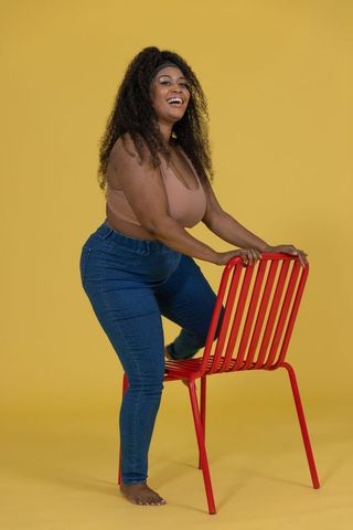 Curvy african american woman laughing leaning on red chair barefoot on yellow backdrop