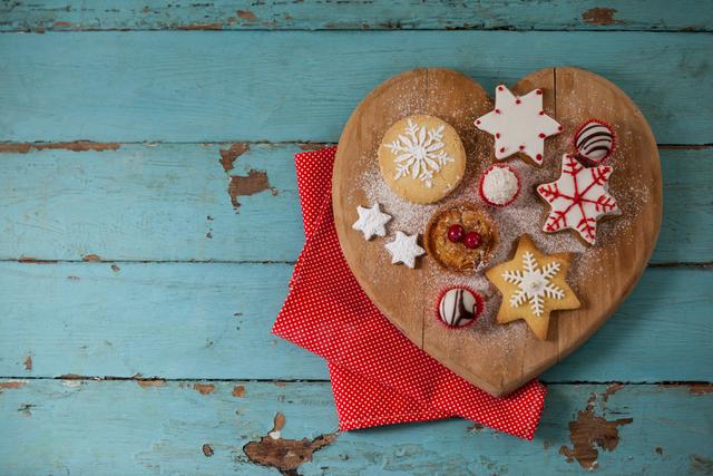Heart-shaped wooden board with assorted Christmas gingerbread cookies decorated with icing, placed on rustic blue wooden table. Red polka dot napkin adds a festive touch. Ideal for holiday-themed content, baking blogs, festive greeting cards, and seasonal advertisements.