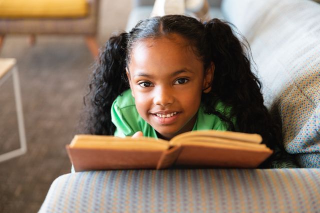 Young African American girl smiling while reading book, lying on sofa in play room. Perfect for educational materials, childhood literacy campaigns, school advertisements, and websites promoting children's activities and learning.