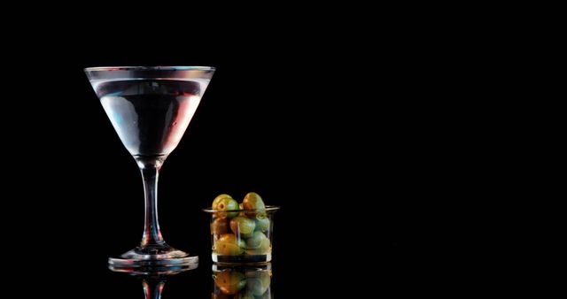 Image showing a martini cocktail in a classic glass with a small dish of green olives against a black background. Ideal for advertisements, bar menus, cocktail parties, nightlife promotions, and drinks recipe blogs. Emphasizes luxury, elegance, and sophisticated lifestyle.