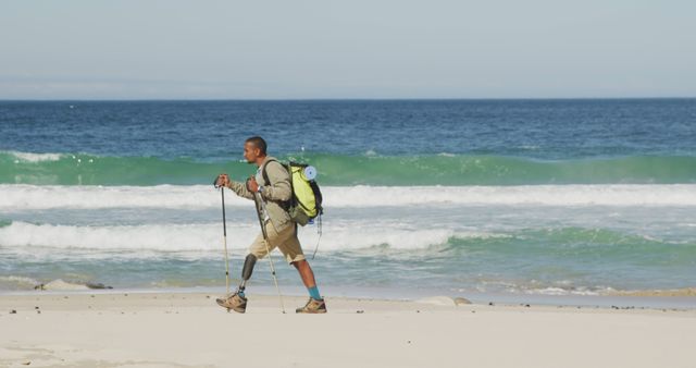 Biracial man with prosthetic leg trekking with backpack and walking poles on a beach by the sea. Long distance walking, fitness, challenge, disability, nature and healthy outdoor lifestyle.