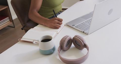 African american woman writing in notebook at tidy desk with laptop coffee and headphones