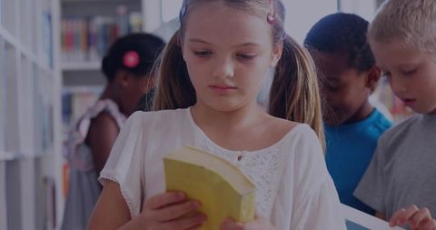 Girl reading paperback in school library with diverse classmates and colorful shelves
